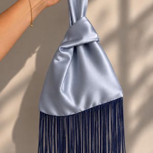 May include: A light blue satin handbag featuring a knotted top handle design and a long, dark blue fringe trim along the bottom edge. The bag has a smooth, reflective surface and a triangular shape created by the knot at the top of the handle.
