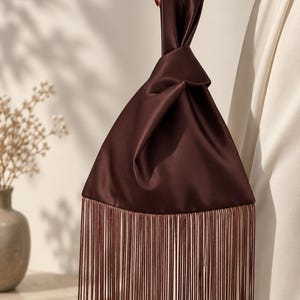May include: A dark brown satin handbag featuring a knotted top handle design and a long, straight fringe trim along the bottom edge. The bag has a smooth, lustrous finish and a triangular shape, with the fringe extending from the base.