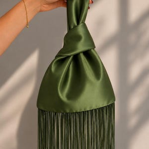 May include: An olive green satin handbag featuring a knotted handle design and a long, straight fringe trim along the bottom edge. The fabric has a smooth, lustrous finish, and the bag is constructed with a single continuous piece of material.