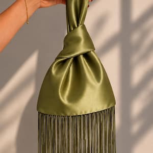 May include: An olive green satin handbag featuring a knotted top handle design and a long, straight fringe trim along the bottom edge. The fabric has a smooth, lustrous finish that reflects light, creating soft folds throughout the body of the bag.