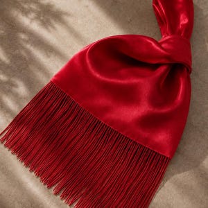 May include: A vibrant red satin handbag featuring a knotted handle design and a long, flowing fringe trim along the bottom edge. The smooth, lustrous fabric creates soft folds and highlights, while the fringe adds texture to the accessory.