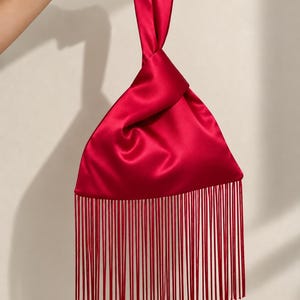 May include: A vibrant red satin handbag featuring a triangular silhouette with a knotted top handle. The bottom edge of the bag is adorned with a long, dense row of matching red fringe that hangs straight down, creating a sleek and polished appearance.