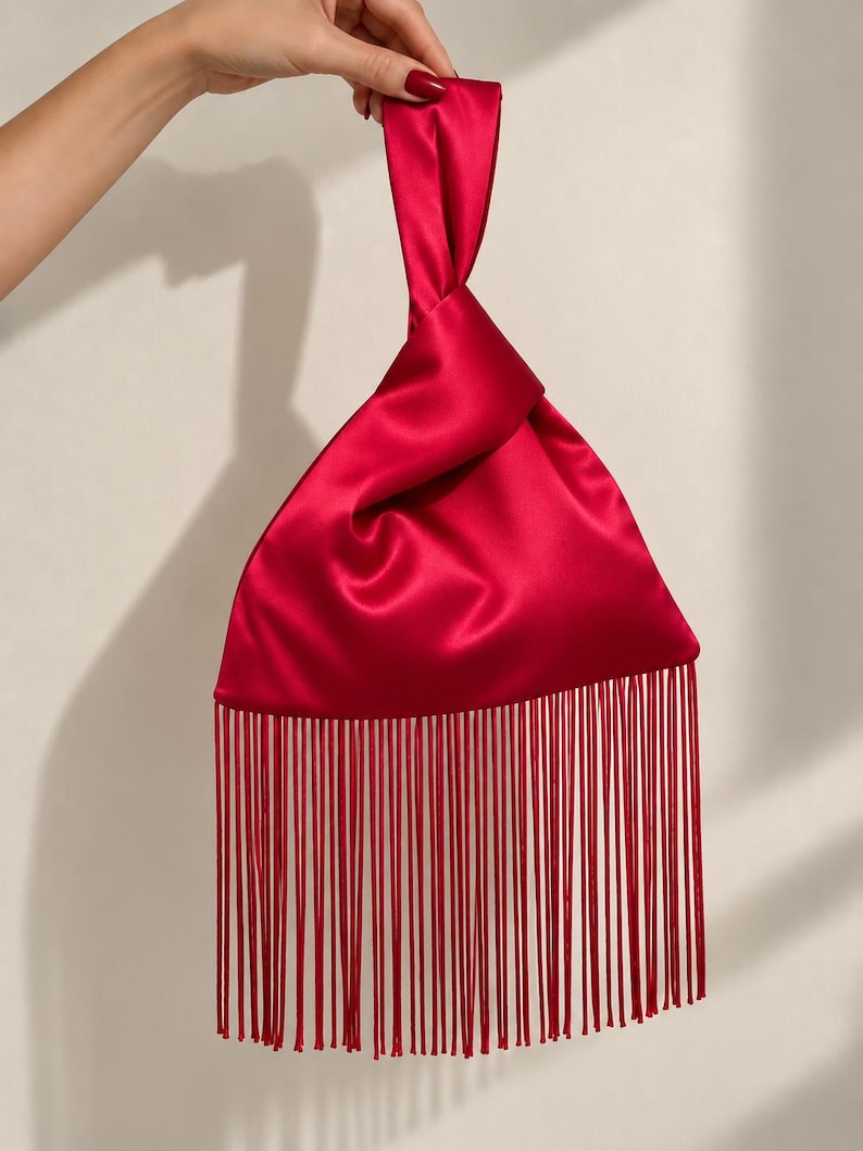 May include: A vibrant red satin handbag featuring a triangular silhouette with a knotted top handle. The bottom edge of the bag is adorned with a long, dense row of matching red fringe that hangs straight down, creating a sleek and polished appearance.