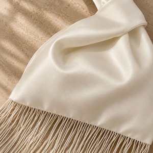 May include: A cream-colored satin handbag featuring a smooth, lustrous finish and a knotted handle design. The bottom edge of the bag is adorned with a long, dense fringe made of thin, matching cream-colored threads that hang vertically downward.