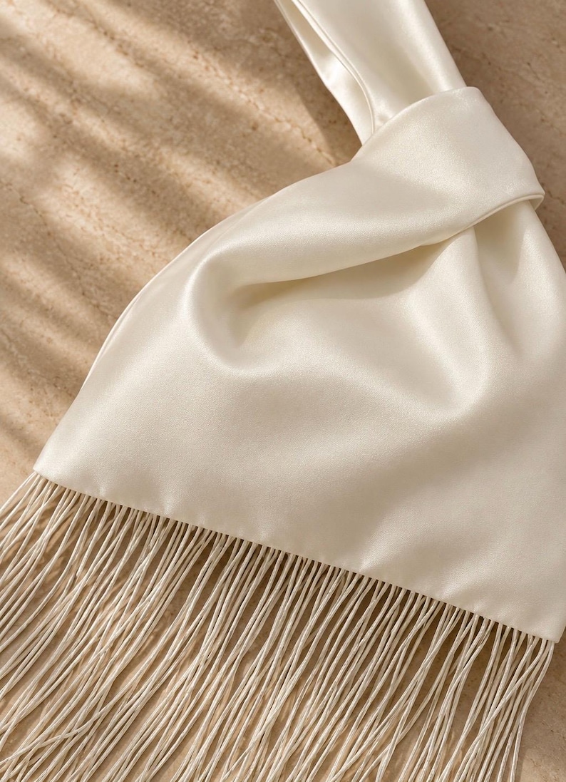 May include: A cream-colored satin handbag featuring a smooth, lustrous finish and a knotted handle design. The bottom edge of the bag is adorned with a long, dense fringe made of thin, matching cream-colored threads that hang vertically downward.