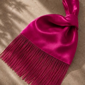 Könnte beinhalten: Eine leuchtend fuchsiafarbene Satin-Handtasche mit einem geknoteten Griffdesign und einem langen, fließenden Fransenbesatz, der von der Unterkante herabhängt. Der Stoff hat einen glatten, reflektierenden Glanz und eine weiche Struktur.