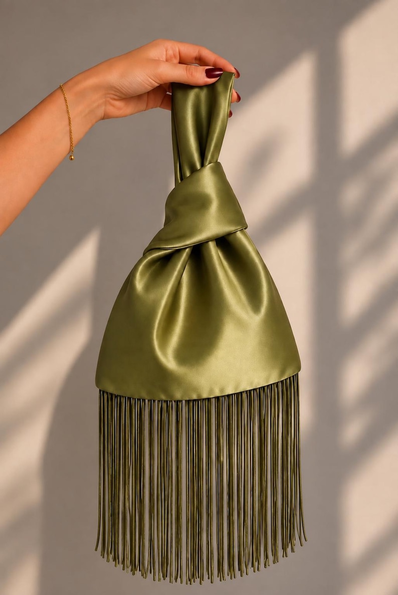 May include: An olive green satin handbag featuring a knotted top handle design and a long, straight fringe trim along the bottom edge. The fabric has a smooth, lustrous finish that reflects light, creating soft folds throughout the body of the bag.