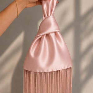 May include: A light pink satin handbag featuring a knotted handle design and a long, straight fringe trim along the bottom edge. The fabric has a smooth, reflective finish, and the bag is constructed with a gathered top section.