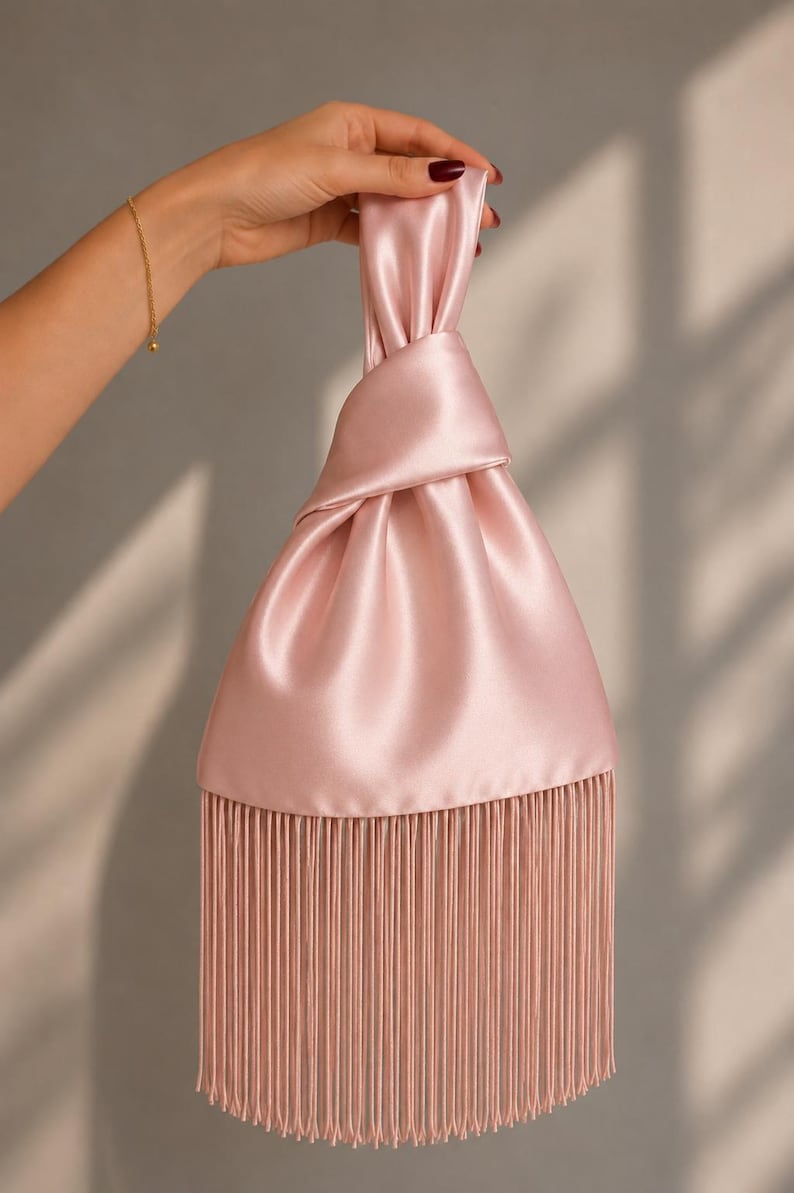 May include: A light pink satin handbag featuring a knotted handle design and a long, straight fringe trim along the bottom edge. The fabric has a smooth, reflective finish, and the bag is constructed with a gathered top section.