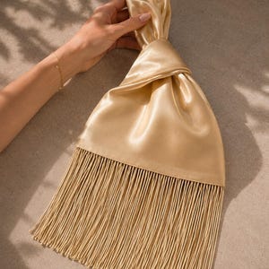 May include: A gold satin handbag featuring a knotted top handle design and a long, flowing fringe trim that extends from the base of the bag. The fabric has a smooth, reflective sheen, and the fringe consists of numerous thin, vertical strands.