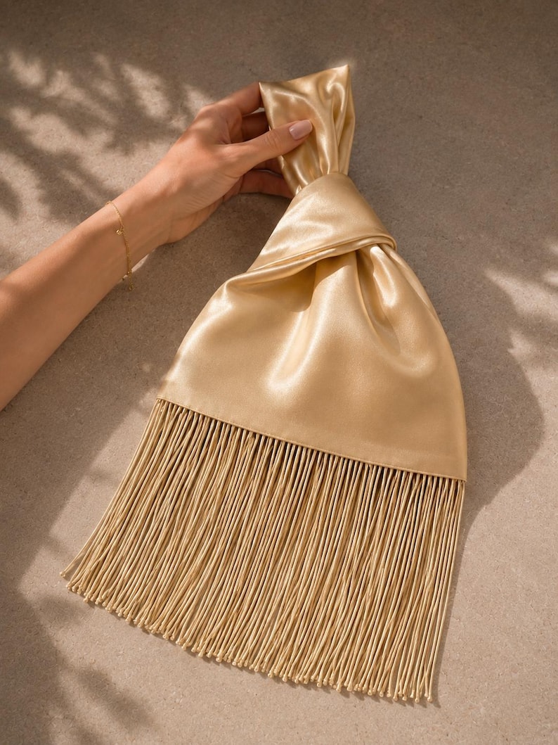 May include: A gold satin handbag featuring a knotted top handle design and a long, flowing fringe trim that extends from the base of the bag. The fabric has a smooth, reflective sheen, and the fringe consists of numerous thin, vertical strands.