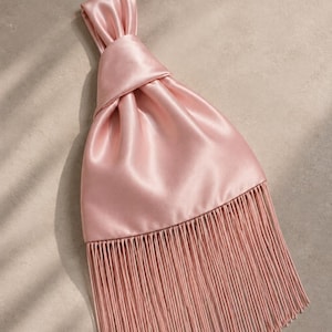 May include: A dusty pink satin handbag featuring a knotted top handle design and a long, straight fringe trim along the bottom edge. The smooth, lustrous fabric creates soft folds leading up to the gathered knot at the top of the bag.