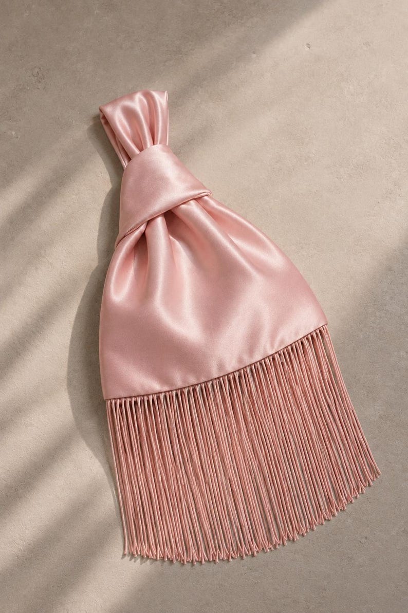May include: A dusty pink satin handbag featuring a knotted top handle design and a long, straight fringe trim along the bottom edge. The smooth, lustrous fabric creates soft folds leading up to the gathered knot at the top of the bag.
