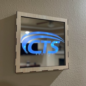 May include: Square mirror with a light wood frame. The mirror features a blue illuminated logo that reads "CTS Designs". The logo is centered on the mirror's surface, and the frame is mounted on a beige wall.