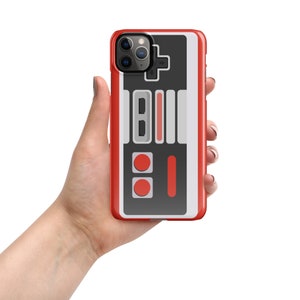 Puede incluir: Carcasa de teléfono roja, blanca y negra con un diseño de mando de videojuego retro. La carcasa tiene un diseño de botones en relieve y un recorte para la cámara.