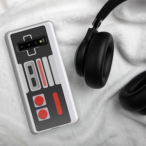 Puede incluir: Una funda de teléfono blanca con un diseño de mando de Nintendo negro y gris. La funda tiene un recorte para la lente de la cámara negra y un diseño de botones rojo y gris.