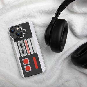 May include: A white and black iPhone case with a retro video game controller design. The case has red buttons and a gray control pad. The case is lying on a white fuzzy surface next to a pair of black headphones.