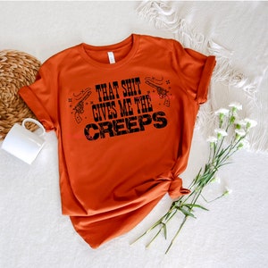 Puede incluir: Una camiseta de color óxido con el texto "That Shit Gives Me The Creeps" en negro. El texto está estilizado con una fuente desgastada e incluye dos pequeñas ilustraciones de pistolas.
