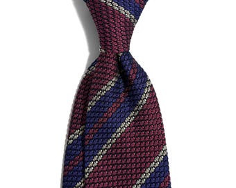 May include: A close-up of a necktie with a textured pattern. The tie features alternating stripes of burgundy, navy blue, and silver. The tie is knotted at the top, showcasing the detailed weave and color variations.