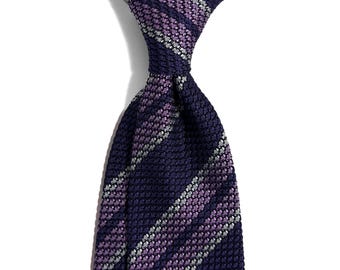 Puede incluir: Una corbata azul marino con un patrón de punto texturizado. La corbata presenta rayas diagonales en tonos morados y plateados. La corbata está anudada en la parte superior, mostrando el tejido y las variaciones de color de la tela. El fondo es blanco liso.
