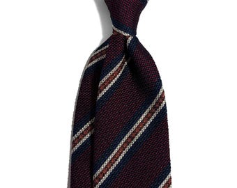 May include: A close-up of a burgundy tie with navy blue and off-white stripes. The tie is knotted, showcasing the textured fabric and color pattern. The background is plain white, highlighting the tie's design.