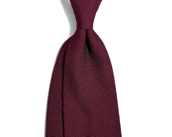 Puede incluir: Primer plano de una corbata burdeos anudada en la parte superior. La corbata tiene un tejido texturizado y está sobre un fondo blanco. El color es un tono rojo intenso y rico.