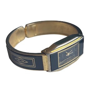 May include: A gold-tone cuff watch featuring a rectangular black face with gold hands and a quartz movement. The band is a hinged bangle design with black enamel panels decorated with gold-colored geometric diamond and rectangular line patterns.