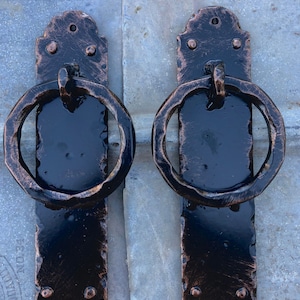 May include: Two black metal door pulls with a ring handle. The pulls have a distressed copper finish and are mounted on a gray background.