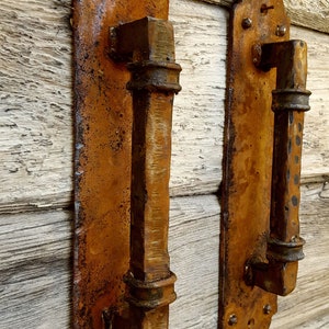 May include: Two rusty metal door handles with a rectangular shape and decorative accents. The handles are mounted on a wooden surface.