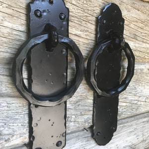 May include: Two black metal door handles with ring pulls. The handles are mounted on a wooden surface.