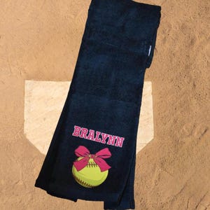 May include: A navy blue softball towel with the name "BRALYNN" in pink lettering. A yellow softball with a pink bow is below the name. The towel is lying on a baseball field.