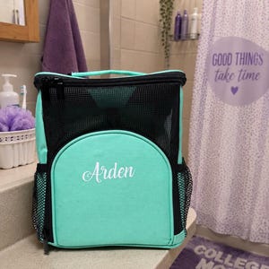 May include: A mint green and black mesh shower caddy with a top handle and side pockets. The front features a rounded pocket with the name Arden embroidered in white script. The bag has a zippered closure and a structured rectangular shape.