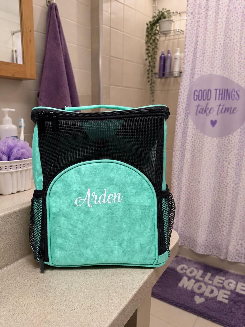May include: A mint green and black mesh shower caddy with a top handle and side pockets. The front features a rounded pocket with the name Arden embroidered in white script. The bag has a zippered closure and a structured rectangular shape.