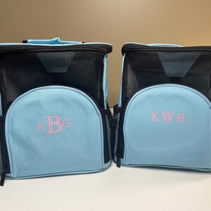 May include: Two light blue backpacks with black mesh panels and black zippers. The left backpack features the embroidered pink monogram ABG, while the right backpack features the embroidered pink monogram KWB. Both bags have side mesh pockets.