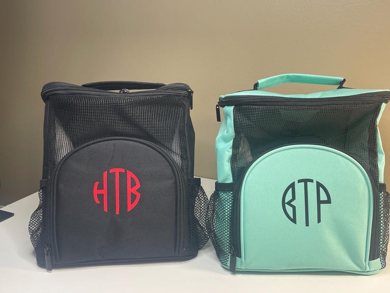 May include: Two square lunch bags with top handles and mesh upper sections. The black bag features a red monogram HTB, while the mint green bag features a black monogram BTP. Both bags have a front zippered compartment and side mesh pockets.