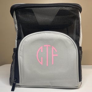 May include: A light gray backpack with black trim and a black mesh top section. The front features a circular monogram with the pink letters GTF. The bag includes black mesh side pockets and a black zipper closure along the top and sides.