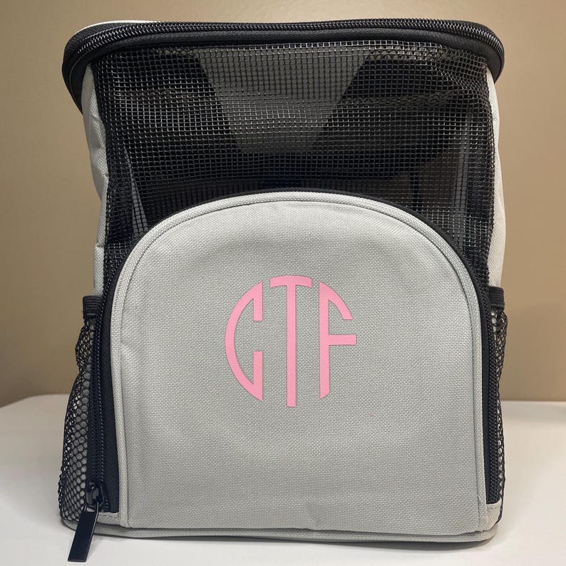 May include: A light gray backpack with black trim and a black mesh top section. The front features a circular monogram with the pink letters GTF. The bag includes black mesh side pockets and a black zipper closure along the top and sides.