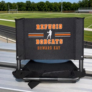 May include: A black stadium seat with a backrest featuring the orange text REFUGIO BOBCATS, a white silhouette of a football player, and the name HOWARD KAY in orange. The seat has a black metal frame and is positioned on silver bleachers.