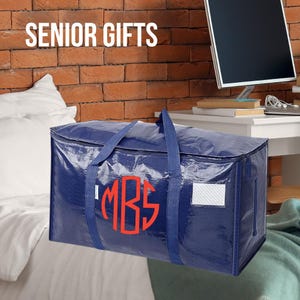 May include: A navy blue storage bag with a red monogram and the text "SENIOR GIFTS" in white. The bag has blue handles and a zipper closure. The bag is sitting on a bed.