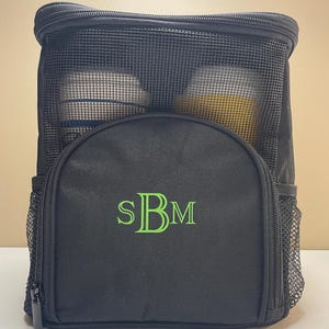 May include: A black rectangular storage bag with a top zipper closure and a mesh upper panel. The front features a semi-circular pocket embroidered with the lime green monogram letters SBM. Two side mesh pockets are attached to the main body.