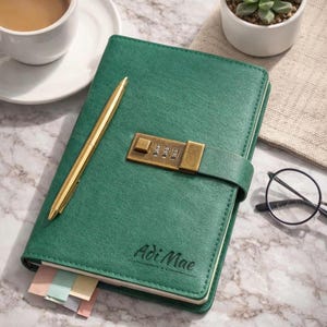 May include: A dark green journal with a gold-colored combination lock and a gold-colored pen resting on the cover. The name Adi Mae is embossed on the front cover. The journal features a strap closure and visible colorful page markers at the bottom.