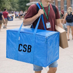 Op de afbeelding: Een grote, rechthoekige blauwe opbergtas met de letters "CSB" in zwart gedrukt. De tas heeft een ritssluiting en twee lange handvatten. De tas wordt door een persoon gedragen.