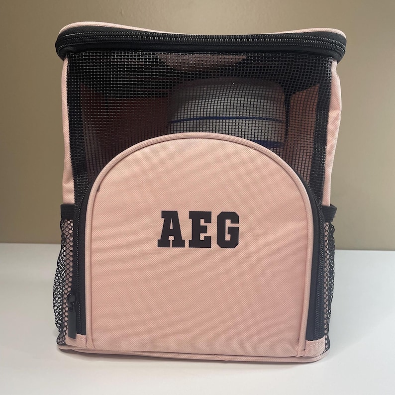 May include: A light pink rectangular storage bag with black trim and black mesh panels on the front and sides. The front features a semi-circular zippered pocket with the letters AEG printed in bold black block lettering in the center.