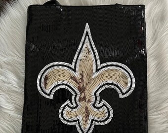 May include: Black tote bag with a gold and white fleur-de-lis design. The bag is made of sequins.