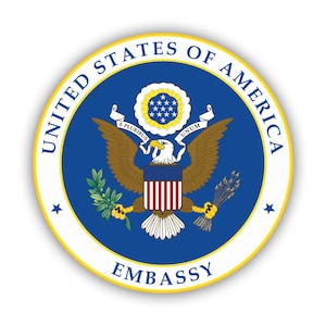 Puede incluir: Un sello azul y blanco con un águila calva sosteniendo un haz de flechas en una garra y una rama de olivo en la otra. El sello está rodeado por un círculo blanco con las palabras "United States of America" y "Embassy" en letras doradas.
