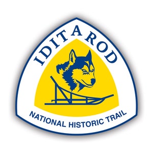 May include: A yellow and blue triangular emblem with the text "Iditarod National Historic Trail" and a stylized image of a husky dog pulling a sled.