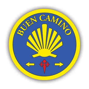 May include: A yellow and blue circular sticker with the text "BUEN CAMINO" in yellow lettering around the edge. A yellow scallop shell is in the center with a red and white cross below it. Two yellow arrows point left and right.