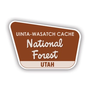 May include: A brown, trapezoidal sticker with a white border featuring the text UINTA-WASATCH CACHE National Forest and UTAH in white and brown lettering. The design mimics a standard road sign with a clean, flat graphic style and rounded corners.