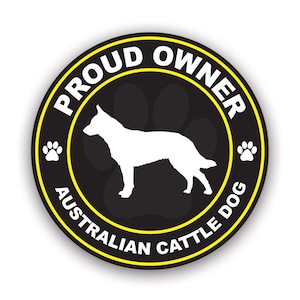 May include: A round black sticker with a yellow border and white text that reads "PROUD OWNER AUSTRALIAN CATTLE DOG." A white silhouette of an Australian Cattle Dog is in the center, with paw prints and text around the edges.