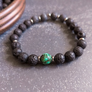 May include: A black lava rock bracelet with a turquoise gemstone bead. The bracelet is made with small, round beads and has a silver metal clasp.
